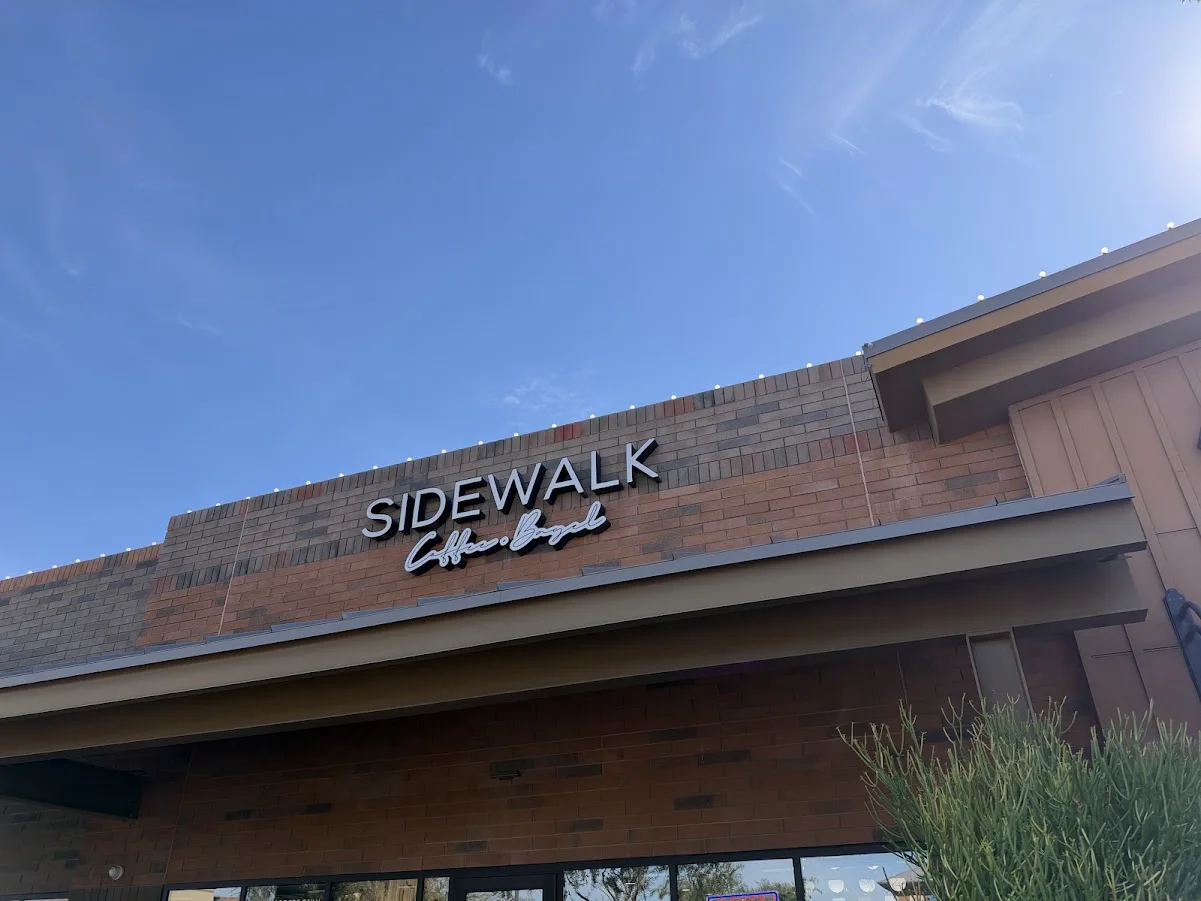 Sidewalk Coffee + Bagel sign on the cafe’s brick facade, viewed from below beneath a blue sky.