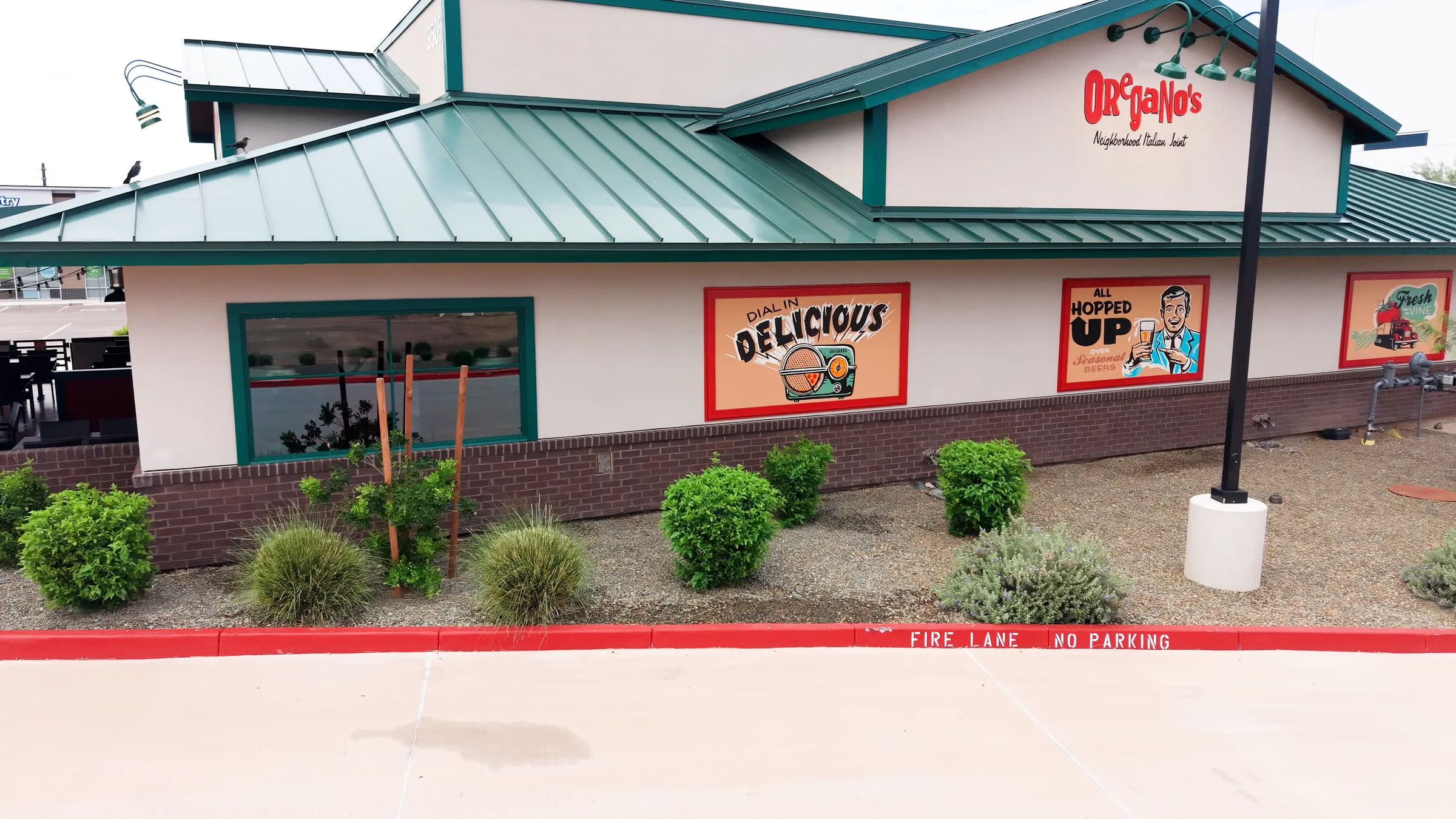 Oregano’s side exterior with red restaurant lettering, green roof and colorful pizza, beer and wine wall panels.