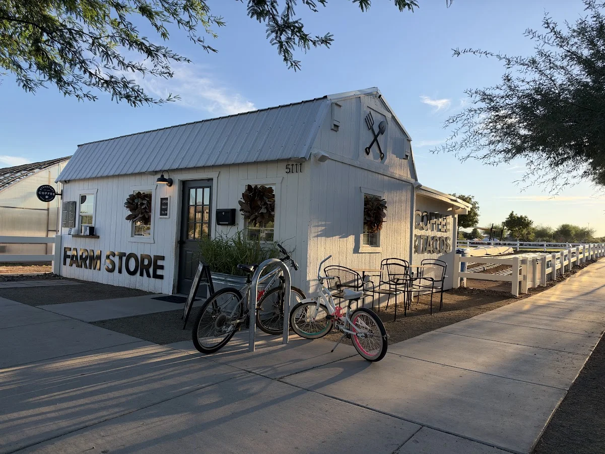 White Steadfast Coffee and Farm Store building at Eastmark, with coffee and tacos signs, outdoor tables and bicycles beside the sidewalk.