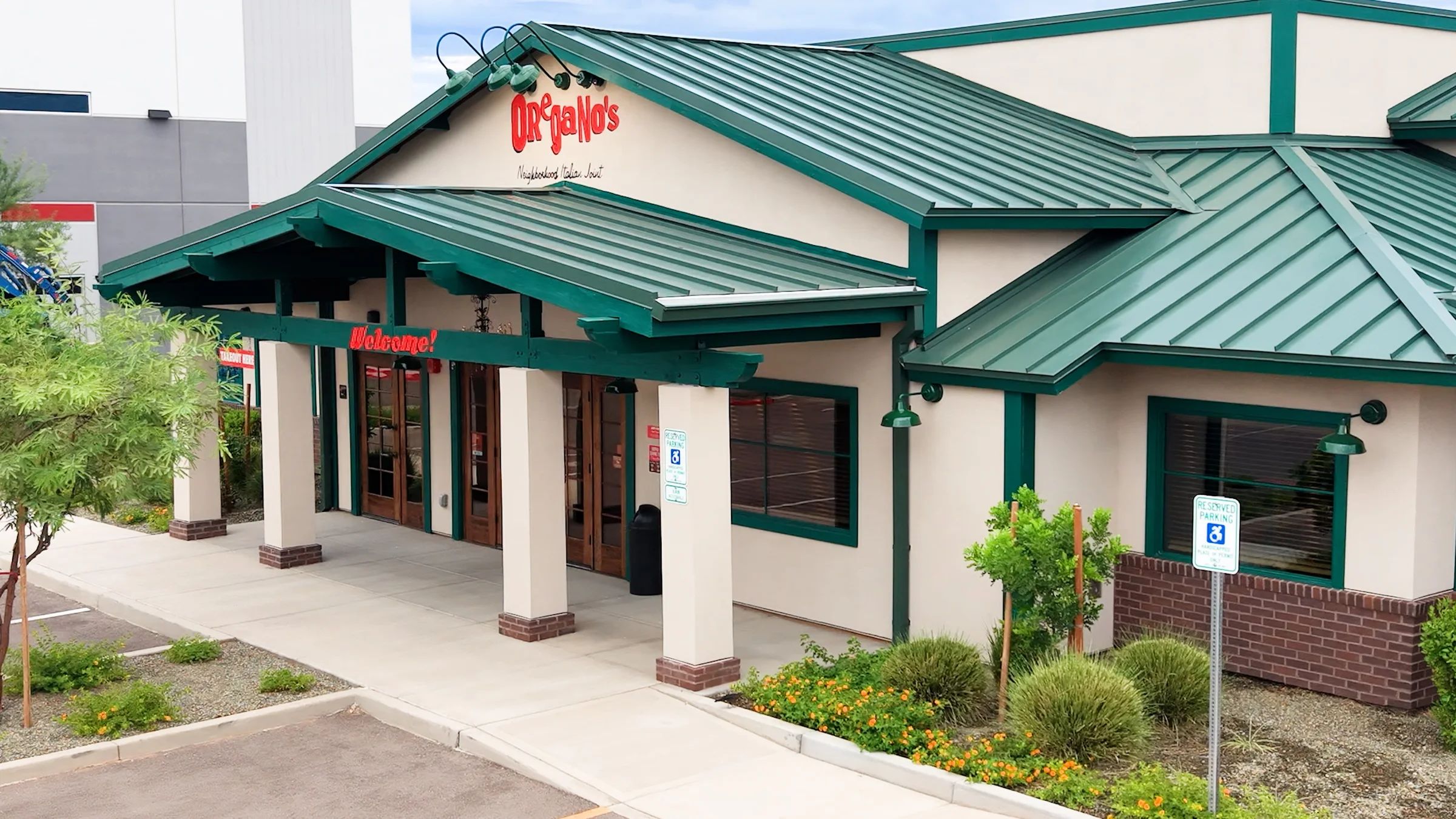 Oregano’s entrance with red lettering, a green metal roof, covered walkway and landscaped parking area in Mesa.