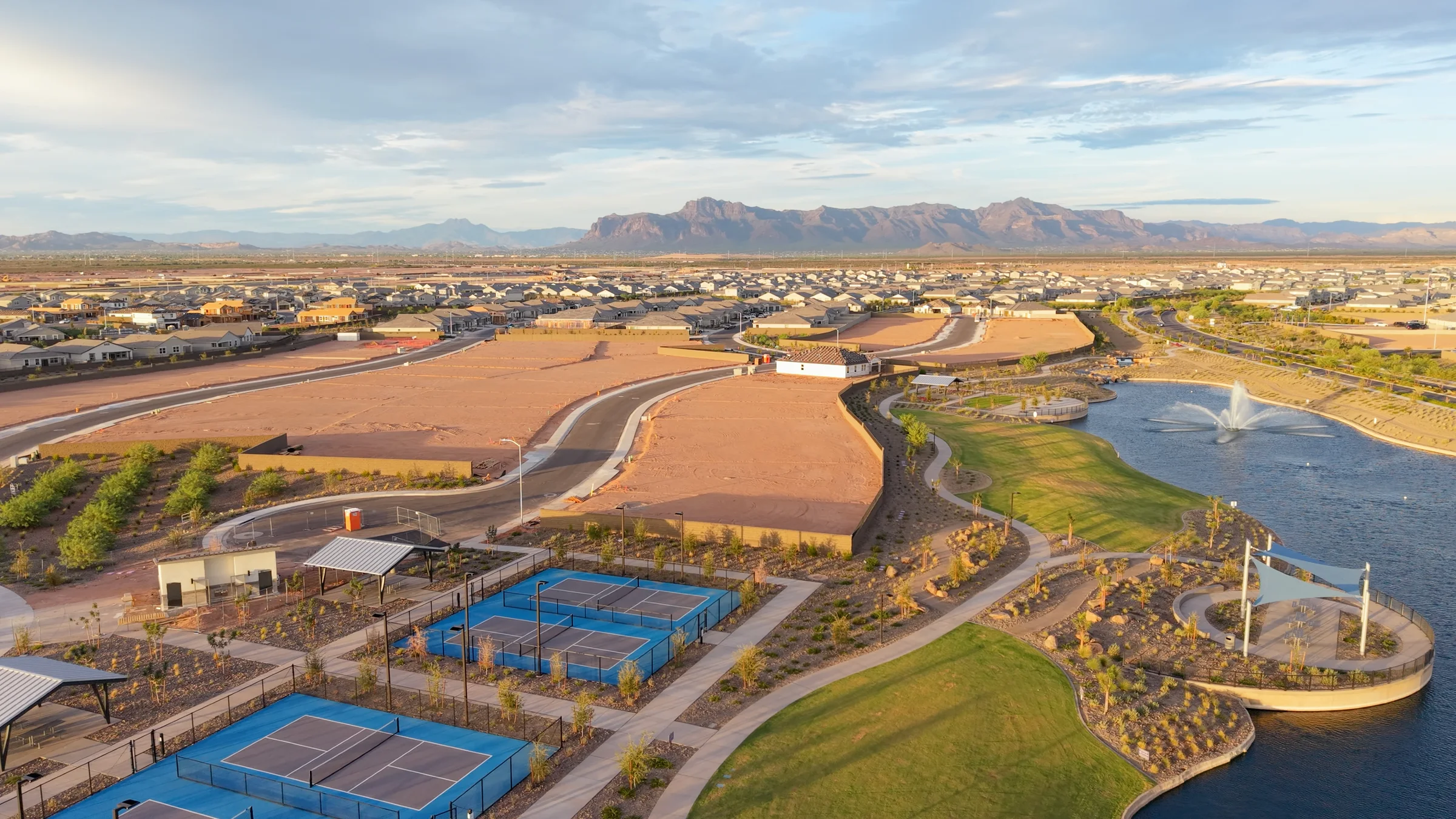 Blue pickleball courts beside Radiance’s lake, shaded gathering spaces and walking paths, with the Superstition Mountains on the horizon.