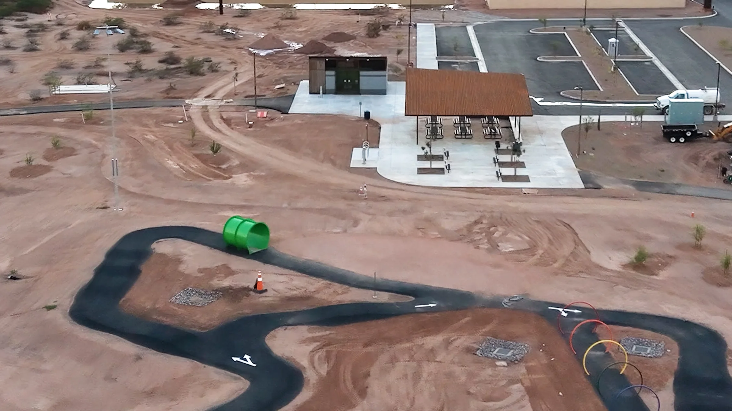 Covered picnic tables, a restroom building and a paved parking lot beside a green tunnel on the new Signal Butte Park bike loop.