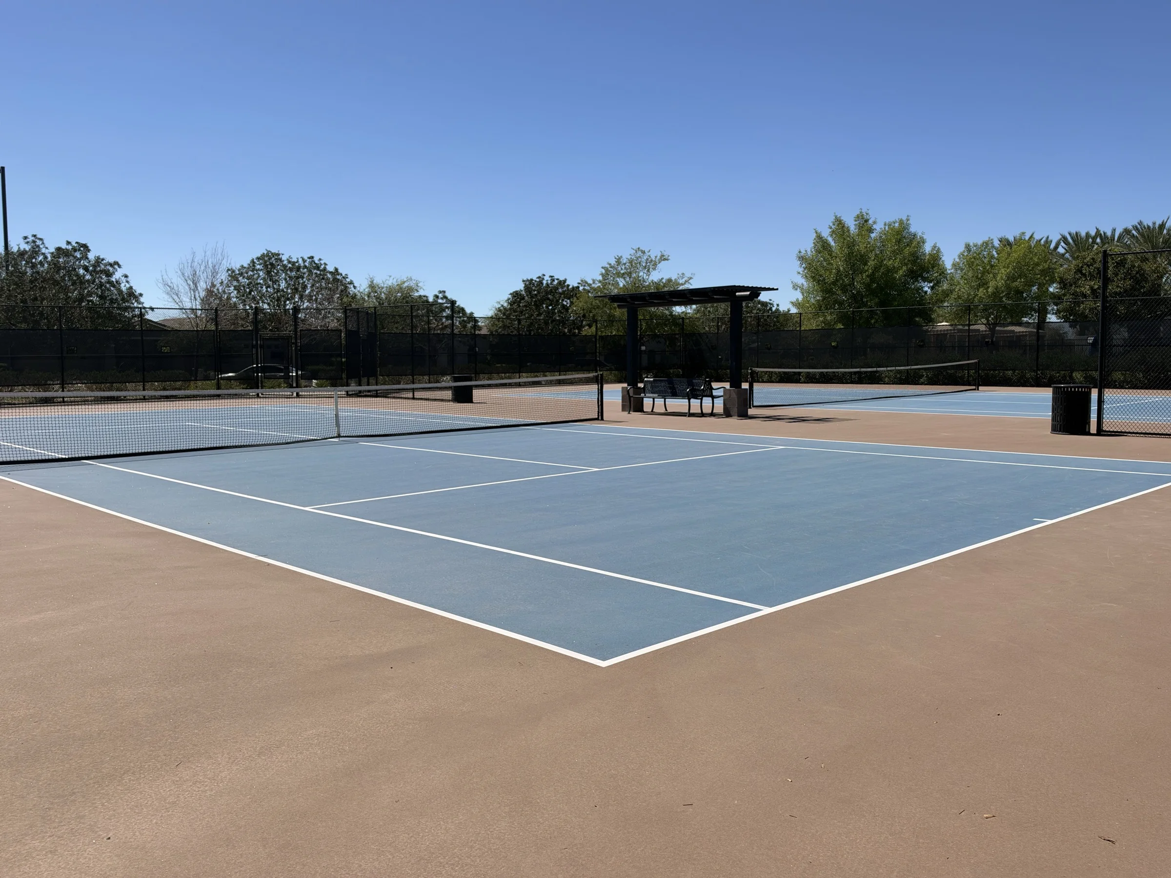 Blue tennis courts with nets and a shaded bench at Cadence at Gateway in Mesa.