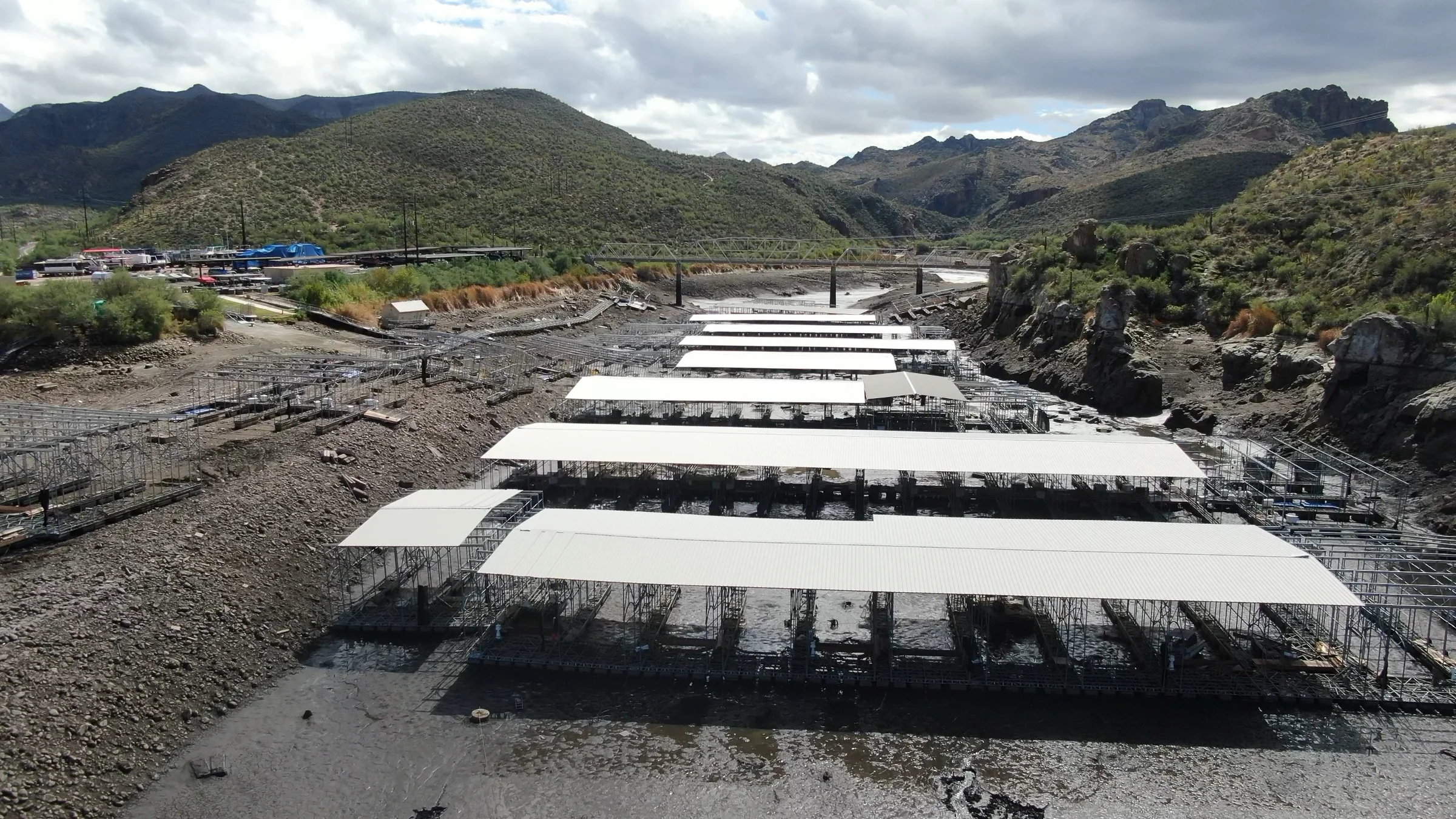 Rows of covered marina structures stand above exposed ground, with a bridge and desert hills behind them at Canyon Lake.