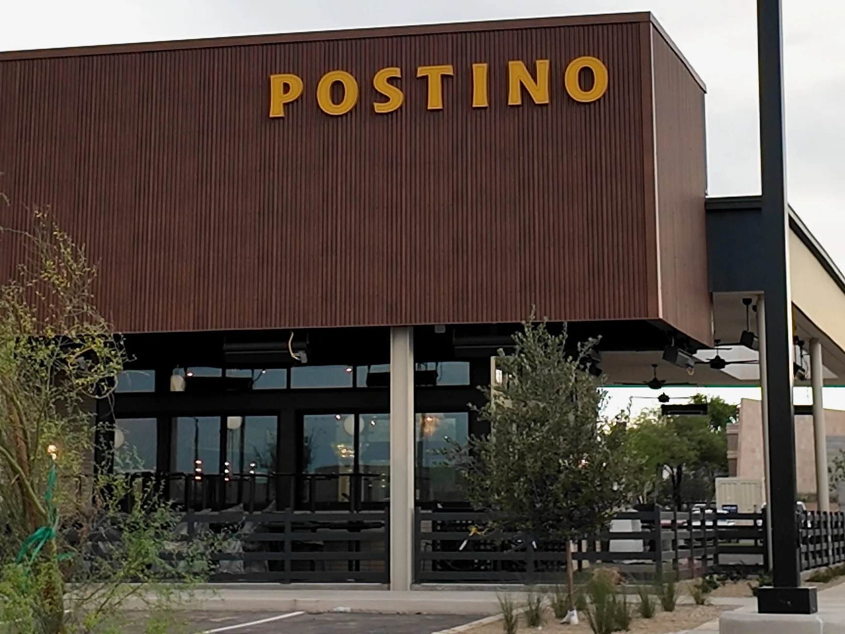 Postino’s gold sign above a covered patio, ceiling fans and a paved walkway.